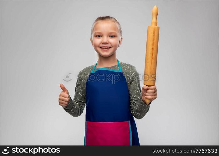 cooking, culinary and baking concept - happy smiling little girl in apron with rolling pin showing thumbs up over grey background. girl in apron with rolling pin showing thumbs up