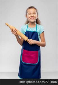 cooking, culinary and baking concept - happy smiling little girl in apron with rolling pin over grey background. smiling little girl in apron with rolling pin