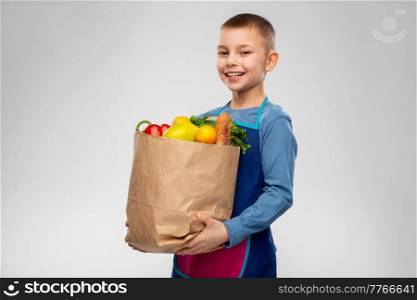 cooking, culinary and eating concept - happy smiling little boy in apron holding paper bag with food over grey background. smiling boy in apron holding paper bag with food