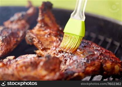 cooking, culinary and food concept - close up of brush smearing marinade sauce on barbecue meat roasting on brazier grill outdoors. close up of barbecue meat roasting on grill