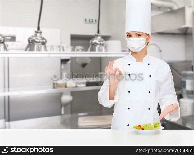 cooking, culinary and health concept - female chef in toque wearing face protective medical mask with food on plate showing ok hand sign over restaurant kitchen background. female chef in mask showing ok sign at kitchen