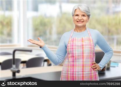 cooking, culinary and old people concept - portrait of smiling senior woman in apron holding something on her empty hand over home kitchen on background. portrait of smiling senior woman in apron at home
