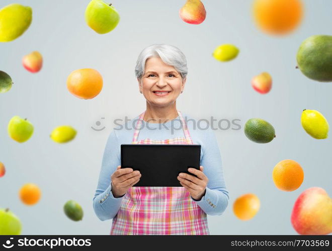 cooking, culinary and old people concept - portrait of smiling senior woman r in kitchen apron with tablet pc compute over fruits on grey background. smiling senior woman in apron with tablet computer