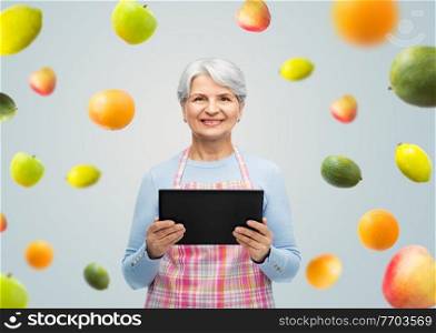 cooking, culinary and old people concept - portrait of smiling senior woman r in kitchen apron with tablet pc compute over fruits on grey background. smiling senior woman in apron with tablet computer