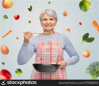 cooking, culinary and old people concept - portrait of smiling senior woman in kitchen apron with frying pan and spoon tasting food over fruits on grey background. smiling senior woman in apron with frying pan