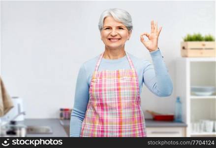 cooking, culinary and old people concept - portrait of smiling senior woman in apron showing ok hand sign over home kitchen background. smiling senior woman showing ok gesture at kitchen