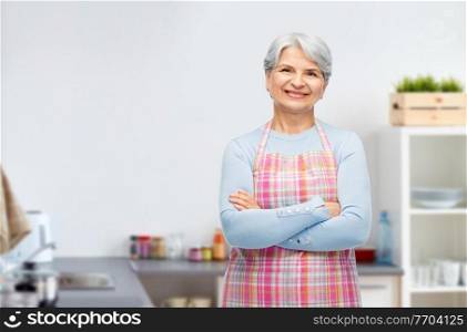 cooking, culinary and old people concept - portrait of smiling senior woman in apron with crossed arms over home kitchen background. portrait of smiling senior woman at kitchen