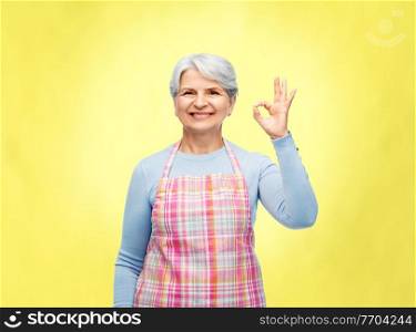 cooking, culinary and old people concept - portrait of smiling senior woman in kitchen apron showing ok hand sign over illuminating yellow background. smiling senior woman in apron showing ok gesture