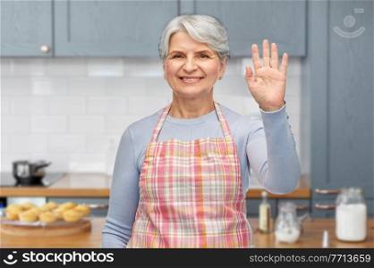 cooking, culinary and old people concept - portrait of smiling senior woman in apron waving hand over home kitchen background. smiling senior woman in apron waving hand