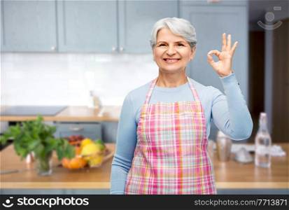 cooking, culinary and old people concept - portrait of smiling senior woman in apron showing ok hand sign over kitchen background. smiling senior woman in apron showing ok gesture
