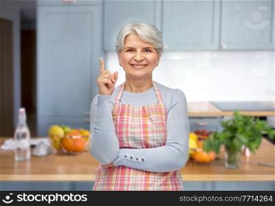 cooking, culinary and old people concept - portrait of smiling senior woman in apron pointing finger up over home kitchen background. smiling senior woman in apron pointing finger up