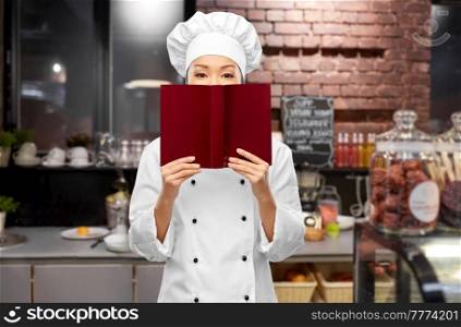 cooking, culinary and people concept - female chef hiding behind cook book over restaurant kitchen background. female chef hiding behind cook book