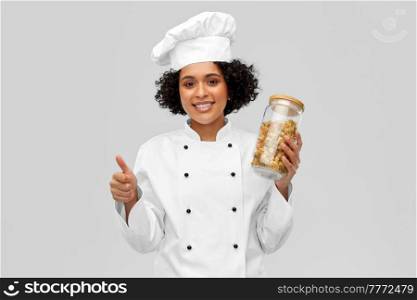 cooking, culinary and people concept - happy smiling female chef holding jar with pasta showing thumbs up over grey background. female chef with pasta in jar showing thumbs up