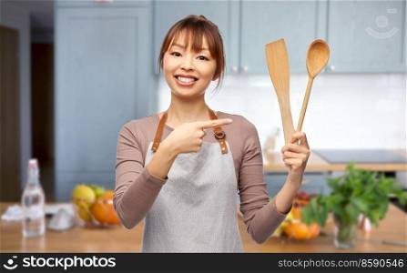 cooking, culinary and people concept - happy smiling female chef in apron with wooden spoon over home kitchen background. woman in apron with spoon and spatula in kitchen