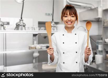 cooking, culinary and people concept - happy smiling female chef in toque with wooden spoon and spatula over restaurant kitchen background. happy smiling female chef with spatulas at kitchen