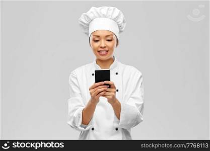 cooking, culinary and people concept - happy smiling female chef in white jacket with smartphone over grey background. smiling female chef with smartphone