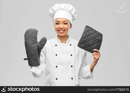 cooking, culinary and people concept - happy smiling female chef in white jacket with potholder and oven mitt over grey background. smiling female chef in jacket with potholders