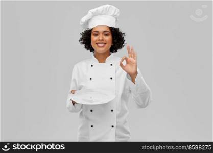 cooking, culinary and people concept - happy smiling female chef in white toque and jacket holding empty plate and showing ok hand sign over grey background. female chef holding plate and showing ok sign