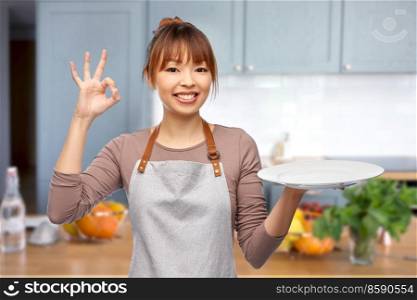 cooking, culinary and people concept - happy smiling female chef or waitress in apron with empty plate over home kitchen background. happy woman in apron with empty plate in kitchen