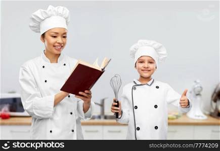 cooking, culinary and people concept - happy smiling female chef reading cook book and little girl holding whisk and showing thumbs up gesture over kitchen background. female chef with cook book and girl in kitchen