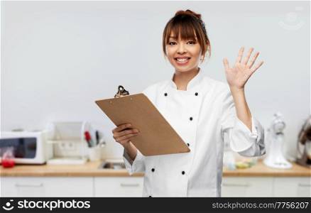 cooking, culinary and people concept - happy smiling female chef with clipboard waving hand over restaurant kitchen background. female chef with clipboard waving hand on kitchen
