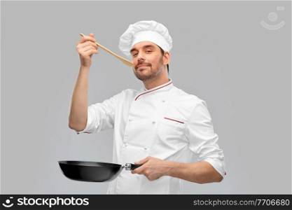 cooking, culinary and people concept - happy smiling male chef in jacket with frying pan and spoon tasting food over grey background. happy male chef with frying pan tasting food