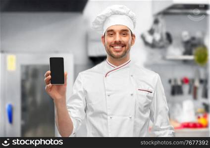 cooking, culinary and people concept - happy smiling male chef in toque showing smartphone over restaurant kitchen background. happy smiling male chef showing smartphone
