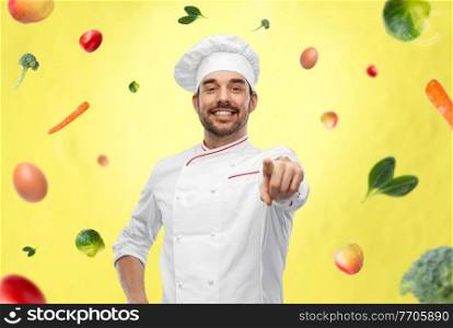 cooking, culinary and people concept - happy smiling male chef in toque pointing finger to camera over vegetables on illuminating yellow background. happy smiling male chef in toque pointing to you