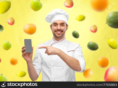 cooking, culinary and people concept - happy smiling male chef in toque showing smartphone over fruits on illuminating yellow background. happy smiling male chef showing smartphone