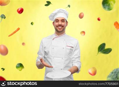 cooking, culinary and people concept - happy smiling male chef in toque holding empty plate over vegetables on illuminating yellow background. happy smiling male chef holding empty plate