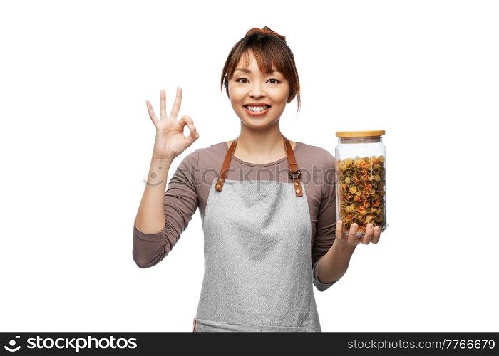 cooking, culinary and people concept - happy smiling woman in apron holding jar with pasta showing ok hand sign over white background. woman in apron with pasta in jar showing ok sign
