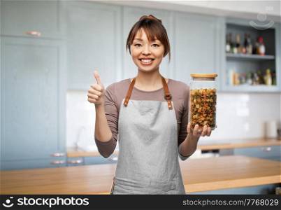 cooking, culinary and people concept - happy smiling woman in apron holding jar with pasta and showing thumbs up gesture over kitchen background. woman in apron with pasta in jar showing thumbs up