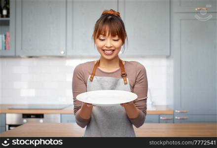 cooking, culinary and people concept - happy smiling woman in apron with empty plate over kitchen background. happy woman in apron with empty plate in kitchen