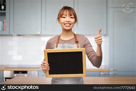 cooking, culinary and people concept - happy smiling woman in apron with chalkboard showing thumbs up gesture over kitchen background. woman in apron with chalkboard showing thumbs up