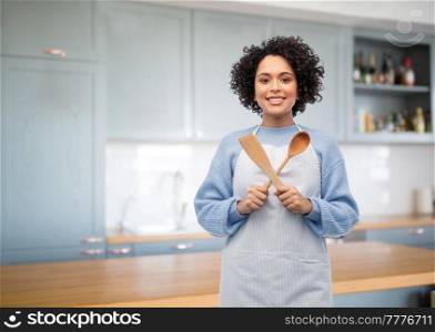 cooking, culinary and people concept - happy smiling woman in apron with wooden spoon and spatula over home kitchen background. woman in apron with spoon and spatula in kitchen