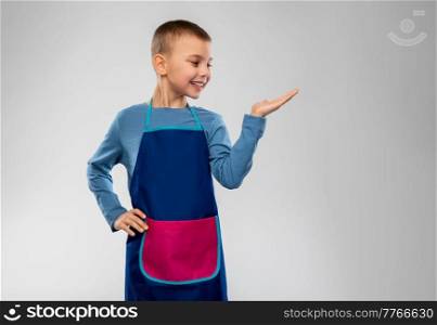cooking, culinary and profession concept - happy smiling little boy in apron holding something imaginary on hand over grey background. little boy in apron holding something on hand
