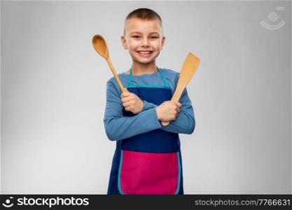 cooking, culinary and profession concept - happy smiling little boy in apron with wooden spoon and spatula over grey background. happy little boy in apron with spoon and spatula