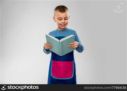 cooking, culinary and profession concept - happy smiling little boy in apron reading cook book over grey background. smiling little boy in apron reading cook book