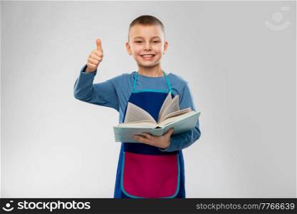 cooking, culinary and profession concept - happy smiling little boy in apron with cook book showing thumbs up over grey background. boy in apron with cook book showing thumbs up