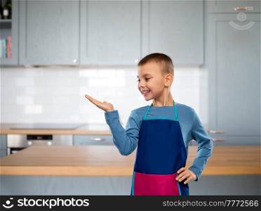 cooking, culinary and profession concept - happy smiling little boy in apron holding something imaginary on hand over kitchen background. little boy in apron holding something on hand
