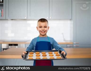 cooking, culinary and profession concept - happy smiling little boy in apron holding baking tray with oatmeal cookies over kitchen background. boy in apron holding baking tray with cookies