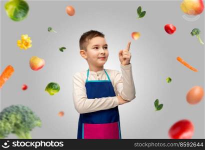 cooking, culinary and profession concept - happy smiling little boy in apron pointing finger up over food on grey background. smiling boy in apron pointing finger up over food