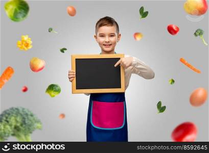 cooking, culinary and profession concept - happy smiling little boy in apron holding chalkboard over food on grey background. little boy in apron holding chalkboard over food