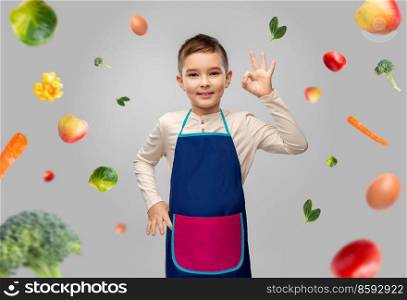 cooking, culinary and profession concept - happy smiling little boy in apron showing ok gesture over food on grey background. smiling little boy in apron over food