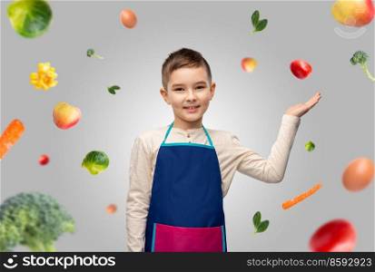 cooking, culinary and profession concept - happy smiling little boy in apron holding something on hand over food on grey background. little boy in apron holding something over food