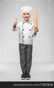 cooking, culinary and profession concept - happy smiling little boy in chef&rsquo;s toque and jacket with rolling pin showing thumbs up over grey background. chef boy with rolling pin showing thumbs up