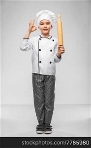 cooking, culinary and profession concept - happy smiling little boy in chef’s toque and jacket with rolling pin showing ok gesture over grey background. boy in chef’s toque with rolling pin showing ok