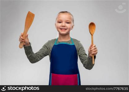 cooking, culinary and profession concept - happy smiling little girl in apron with wooden spoon and spatula over grey background. happy little girl in apron with spoon and spatula
