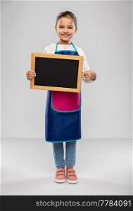 cooking, culinary and profession concept - happy smiling little girl in apron holding chalkboard over grey background. smiling little girl in apron holding chalkboard