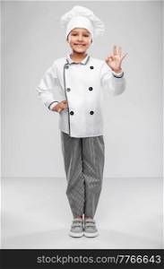 cooking, culinary and profession concept - happy smiling little girl in chef&rsquo;s toque and jacket showing ok gesture over grey background. little girl in chef&rsquo;s toque showing ok gesture
